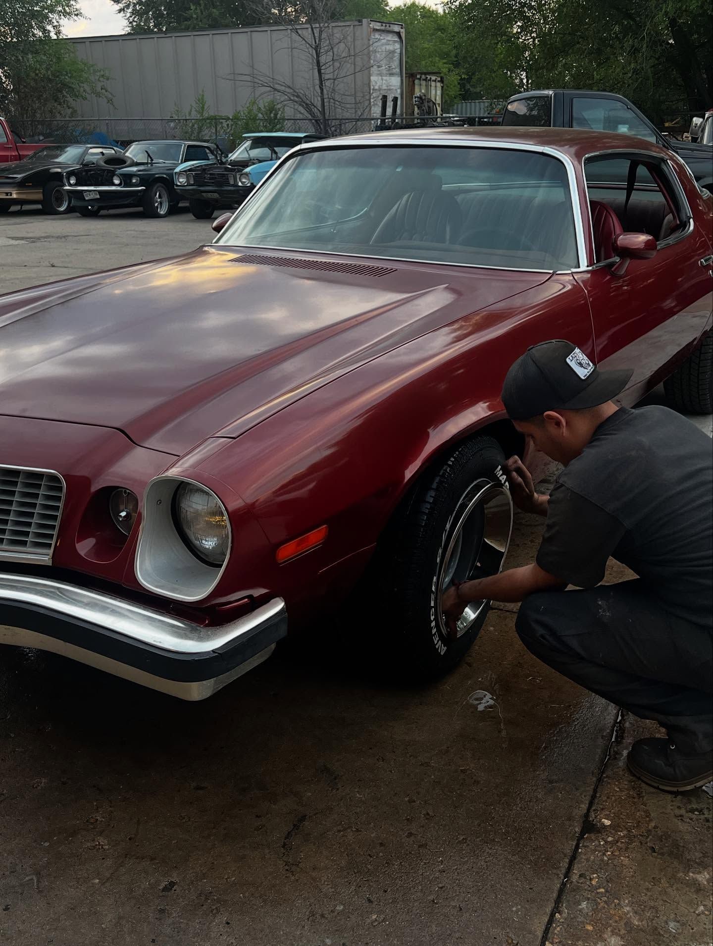 A technician checking the wheel of a maroon classic Camaro, a lot full of classic cars behind it